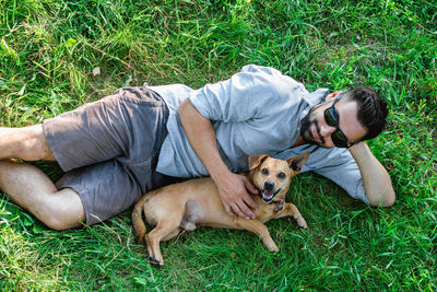 Attractive european man in casual clothing and sunglasses is lying on grass and hugging his dog. 