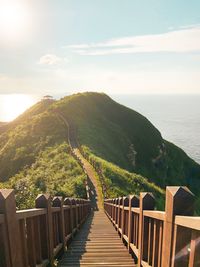 Footbridge leading towards mountains by sea against sky