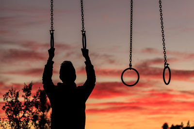 Silhouette man hanging on gymnastic rings against orange sky
