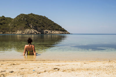 Rear view of woman in bikini sitting on beach against sky