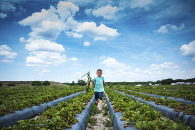 Boy holding root vegetables while running at organic farm against cloudy sky