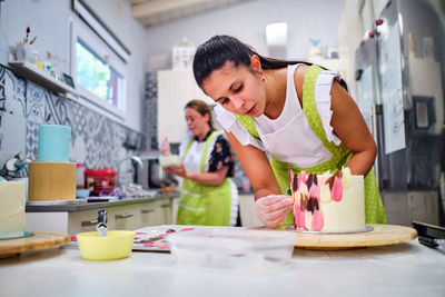 Female confectioner in apron and gloves preparing delicious sweet cake decorated with colorful edible chocolate in light room