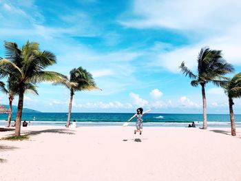 Full length of mid adult woman with arms outstretched jumping at beach against blue sky during sunny day