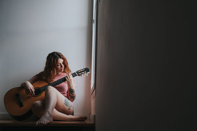 Young red haired laughing woman in pajama with guitar looking at window and sitting on stairs in darkness