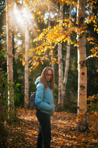 Smiling young woman standing by tree in forest during autumn