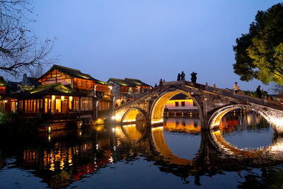 Bridge over river by illuminated buildings against sky