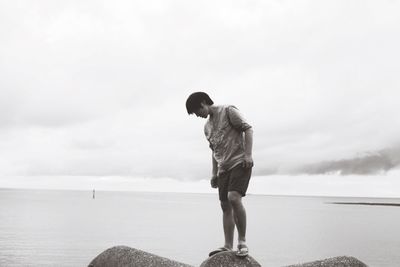 Young man standing on rock at beach against cloudy sky