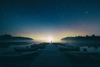 Distant view of person standing on jetty by boats moored on lake against starry sky