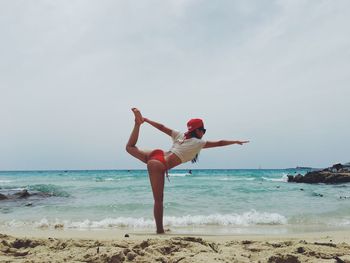 Side view of young woman standing on one leg holding foot at beach against sky