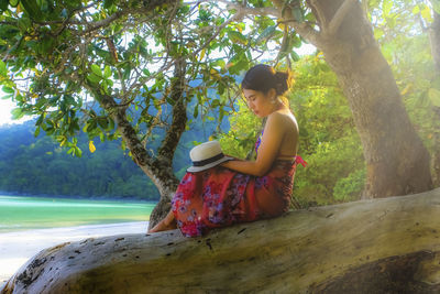 Side view of woman with hat sitting on tree at beach