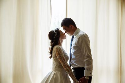 Side view of wedding couple standing face to face by curtain
