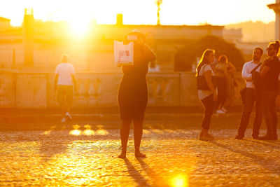 Woman photographing while standing on street during sunset with tourist in background