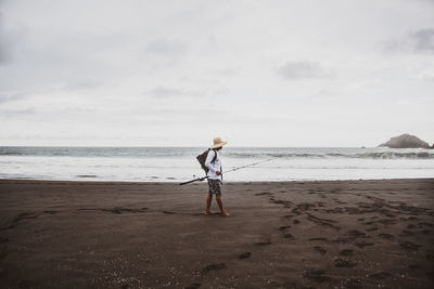 Full body side view of anonymous man with fishing rod with rucksack strolling on sandy shore near sea in nature