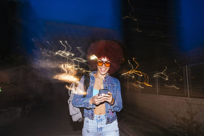 Cheerful female with sunglasses sending text message while standing near fence with light effect in evening time