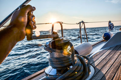 Cropped hand of man rigging rope on boat in sea against sky during sunset