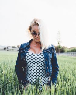 Portrait of young woman wearing denim jacket standing amidst grassy field against clear sky