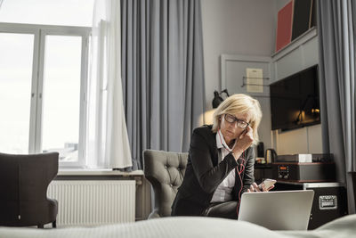 Mature businesswoman adjusting headphones sitting in front of laptop at hotel room