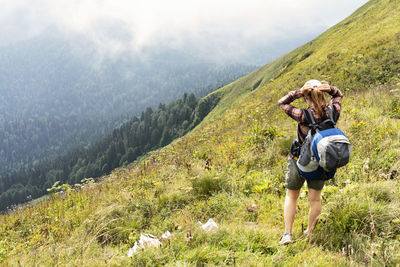 Young woman in plaid shirt with big backpack wearing cap hiking in green mountains in summer 