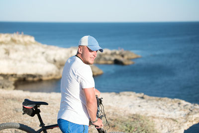 Man with bicycle standing on rock formation at sea against clear sky