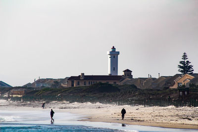 People walking at beach by lighthouse against clear sky