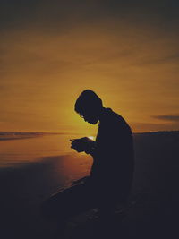 Silhouette man sitting on beach against sky during sunset