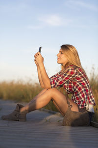 Side view of young woman using mobile phone against sky