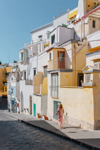 Back view of unrecognizable female in dress walking on asphalt walkway on street near aged buildings with colorful walls in town on procida island in sunny day