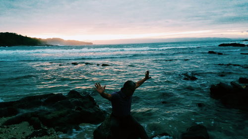 Man with arms outstretched sitting at sea against sky during sunset