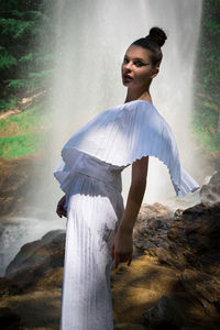Portrait of model wearing white dress posing against waterfall at forest