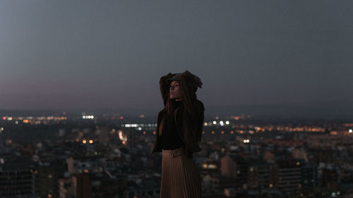 Portrait of woman standing by illuminated cityscape against sky at night