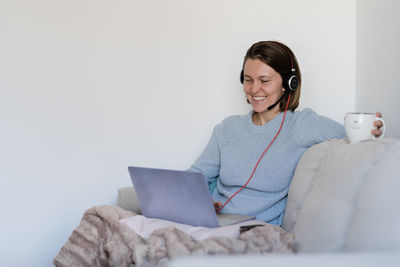 Smiling young woman video conferencing over laptop while sitting on sofa against wall