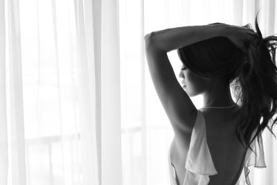 Rear view of young woman standing against window at home