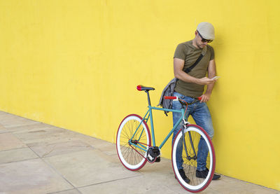 Man using phone while standing with bicycle against yellow wall