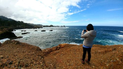 Woman photographing sea while standing on rock formation against sky