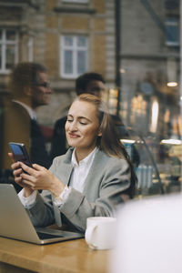 Smiling businesswoman using smart phone in cafe seen through glass window