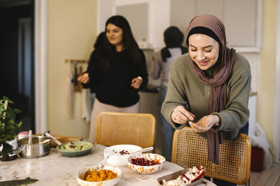 Smiling young woman in hijab garnishing food with pomegranate in kitchen at home