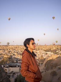 Young man standing against hot air balloons flying in city against sky