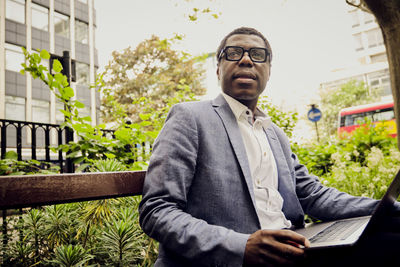 Thoughtful businessman sitting with laptop on bench in park