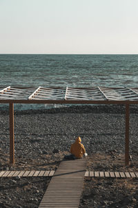 Hooded man sitting alone at end of boardwalk ending at edge of rocky coastal beach