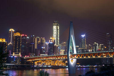 Illuminated bridge over river by buildings against sky at night