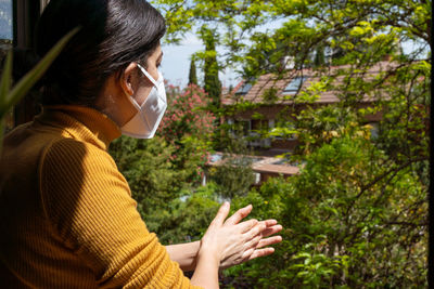 Side view of woman wearing mask standing by window at home