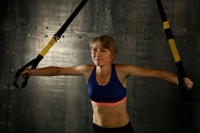 Smiling mature woman pulling cable in gym while standing against wall