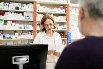 Female pharmacist reading prescription to customer at checkout counter in pharmacy store