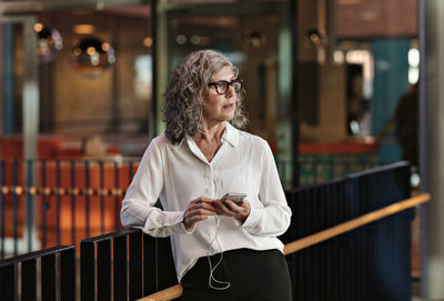 Contemplating senior businesswoman using smart phone while standing at railing in office