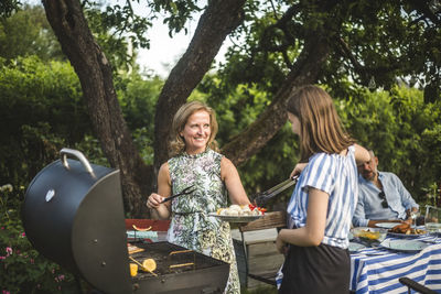 Smiling mother looking at daughter assisting in preparing food at barbecue grill in backyard during weekend party