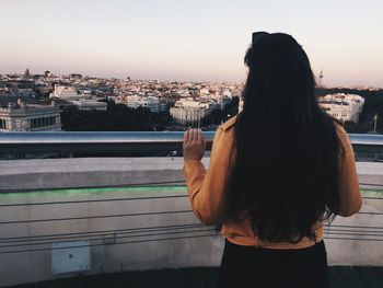 Rear view of woman standing by railing against cityscape during sunset