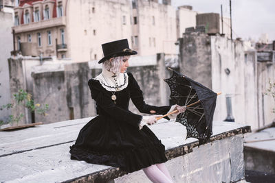 Woman with umbrella wearing hat sitting on rooftop against building