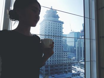 Woman holding coffee cup while standing against window