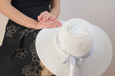 Midsection of woman holding hat while sitting against white wall
