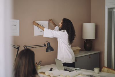 Side view of female architect sticking blueprint on wall while working at office